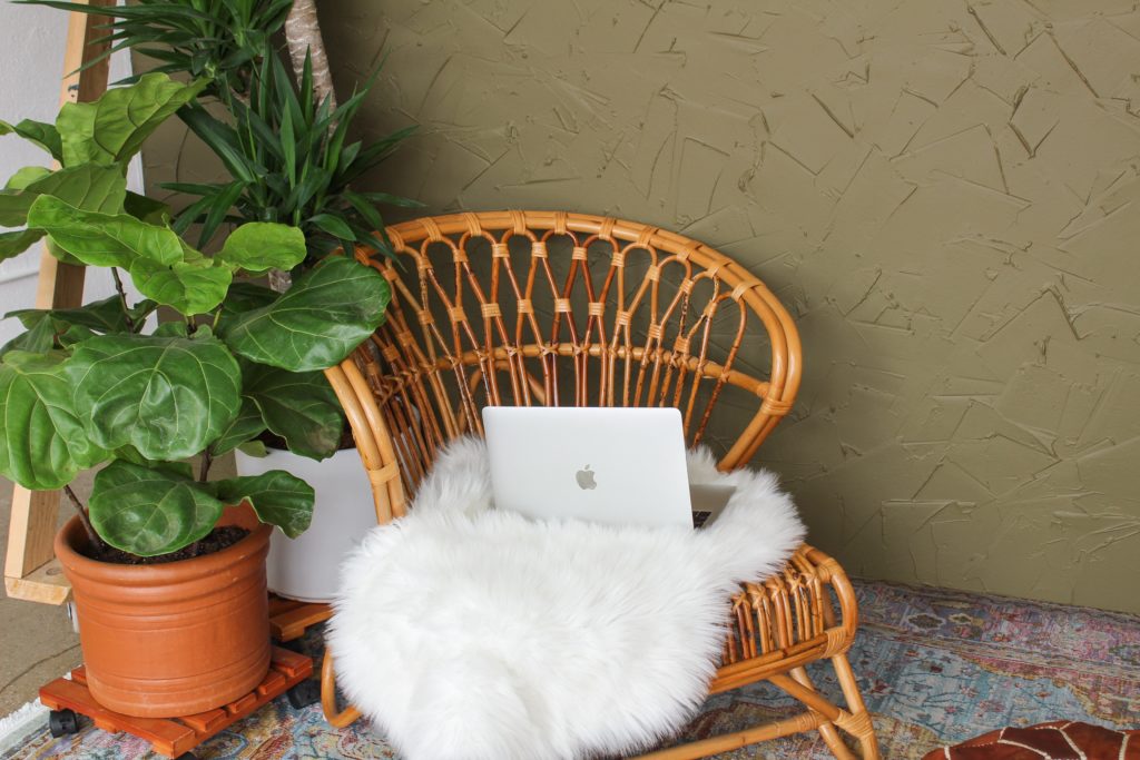 Photograph of rattan chair with an Apple laptop and fluffy throw on it, next to a potted plant