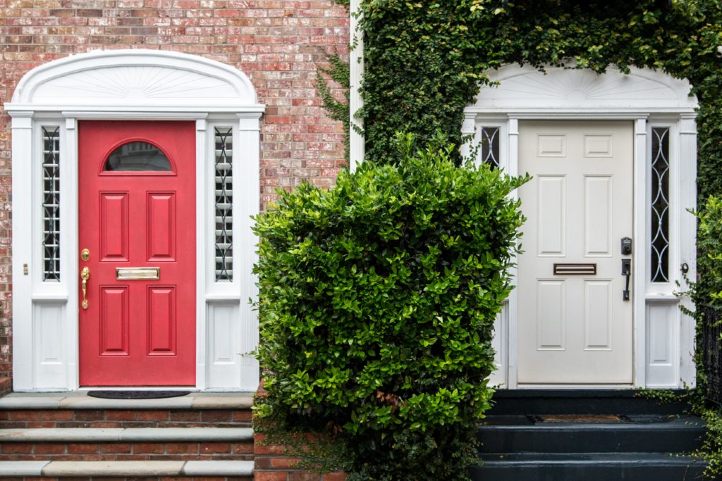 Photograph of two front doors, one red, one cream, with the cream door surrounded by leaves
