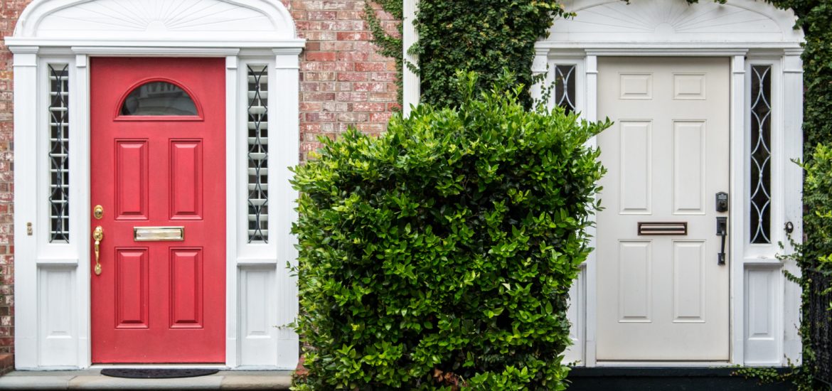 Photograph of two front doors, one red, one cream, with the cream door surrounded by leaves